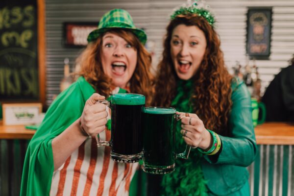 Two women wearing green and drinking green beers.